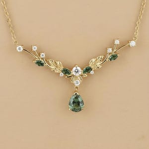 May include: A gold-toned necklace featuring a leaf and floral design. The necklace has a central teardrop-shaped green gemstone pendant, surrounded by white and green gemstones. The chain is delicate and the overall design is elegant.