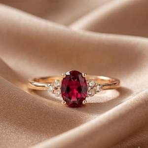 May include: A gold ring featuring a central oval-cut deep red gemstone held by four prongs. The band is accented on each side by a cluster of three small, round, clear white stones set in gold, creating a symmetrical design with a polished finish.