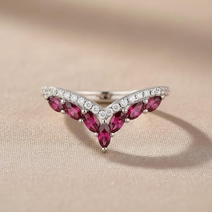 Op de afbeelding: Een zilverkleurige ring met een V-vormig ontwerp. De ring heeft een rij kleine, heldere stenen langs de bovenkant en een reeks marquise-geslepen, dieprode edelstenen in het midden. De ring is geplaatst tegen een neutrale achtergrond.