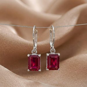 Può includere: Un paio di orecchini pendenti color argento con pietre rettangolari sfaccettate rosso scuro. Ogni orecchino è sospeso a una chiusura a leva, caratterizzato da un design pulito e simmetrico con montature in metallo lucido e gemme rosse.