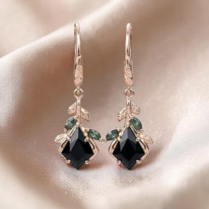 May include: A pair of rose gold earrings with a leaf design. Each earring features a black, square-cut gemstone and small green accent stones. The earrings have a long, slender hook and a decorative leaf and gemstone cluster.
