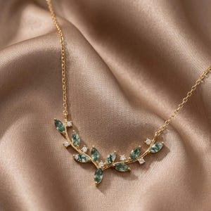 May include: A gold-tone necklace featuring a delicate branch-shaped pendant adorned with marquise-cut moss agate stones and small clear round crystals. The pendant is attached to a thin gold-tone chain that rests in a curved, symmetrical arrangement.