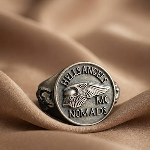 Op de afbeelding: Een cirkelvormige ring van zilverkleurig metaal met een verhoogd embleem van een gevleugelde schedel in het midden. De tekst HELLS ANGELS MC NOMADS is in reliëf aangebracht rond de rand van het cirkelvormige vlak. De band heeft donkere details.