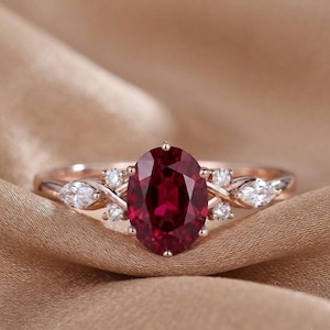 May include: A rose gold ring featuring a large, oval-cut deep red gemstone at the centre. The band is accented on both sides by a cluster of small, clear, round-cut stones and a single marquise-cut stone, creating a delicate, symmetrical design.