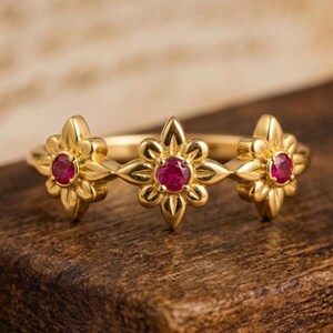 May include: A gold ring featuring three flower-shaped settings, each holding a small, round, red gemstone. The ring is set against a blurred wooden background, highlighting the intricate floral design and the vibrant red of the gemstones.