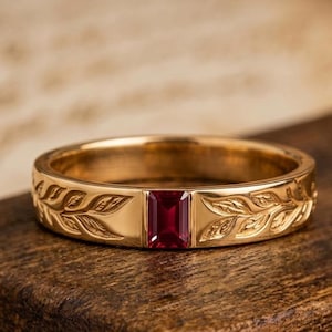 Può includere: Un anello a fascia in oro con una pietra preziosa rosso scuro a taglio rettangolare al centro. La superficie metallica dell'anello è incisa con un dettagliato motivo a foglie che si estende su entrambi i lati della pietra, creando un disegno botanico continuo.