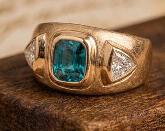 May include: A gold ring featuring a central emerald-cut teal gemstone flanked by two triangular-cut clear stones. The band has a brushed matte finish and a wide, tapered design that holds the three stones in secure, bezel-style settings.