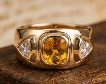 May include: A gold ring featuring a central cushion-cut yellow gemstone set in a bezel, flanked by two triangular-cut clear white gemstones on either side. The band is wide and polished, showcasing a smooth, reflective metallic surface throughout.