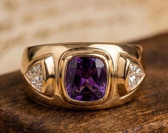 May include: A gold ring featuring a central cushion-cut purple gemstone set in a smooth bezel. The band includes two triangular-cut clear stones, one on each side of the center setting, integrated into the polished gold metal band design.