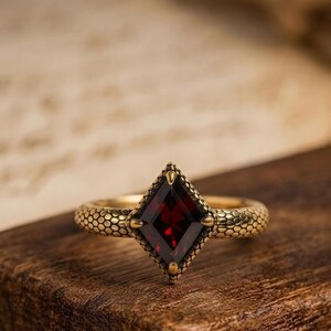 May include: A gold-toned ring featuring a central kite-shaped deep red gemstone. The band and the setting surrounding the stone are textured with a repeating scale pattern, resembling snake skin, with four small prongs securing the gemstone.