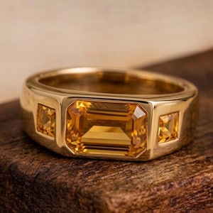Puede incluir: Un anillo de oro con una gema amarilla grande, rectangular y de corte esmeralda en el centro, flanqueada por dos gemas amarillas más pequeñas de corte cuadrado a cada lado. La banda es de oro pulido con una estructura sólida y ancha.
