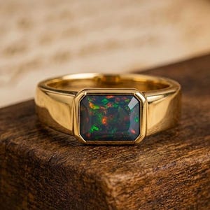Può includere: Un anello in oro con una pietra preziosa opale nera rettangolare sfaccettata. La pietra mostra vivaci bagliori di colore verde e rosso sulla sua base scura. La fascia è in oro liscio e lucido e l'incastonatura trattiene la pietra in una cornice.