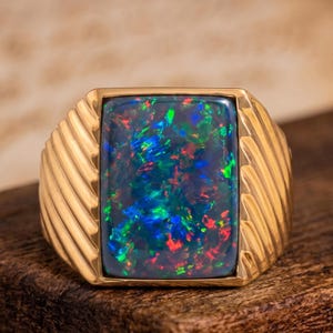 Könnte beinhalten: Ein Goldring mit einem großen, rechteckigen schwarzen Opal, der lebhafte blaue, grüne und rote Lichtreflexe aufweist. Die Ringschiene ist aus poliertem Gold gefertigt und zeigt auf beiden Seiten des Mittelsteins eine diagonale Riffelung.