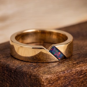 Op de afbeelding: Een gepolijste gouden ring met een enkele rechthoekige zwarte opaal inlay. De opaal vertoont levendige flitsen van rode en groene kleur. De metalen band heeft een glad, reflecterend oppervlak met een diagonale inkeping voor de edelsteen.