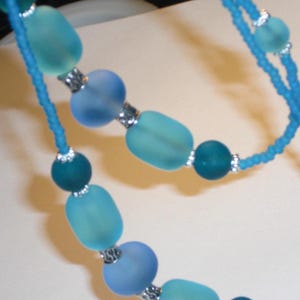 May include: A close-up of a necklace featuring a mix of blue and teal beads. The necklace has a delicate design with various shapes of beads, including round and oblong, connected by a thin, light blue string. Silver-colored accents are visible between the beads.