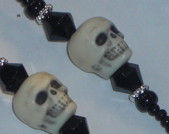 May include: Close-up of a necklace featuring off-white skull beads, black faceted beads, and small silver-colored spacer beads. The skulls have detailed facial features. The necklace is on a light background.