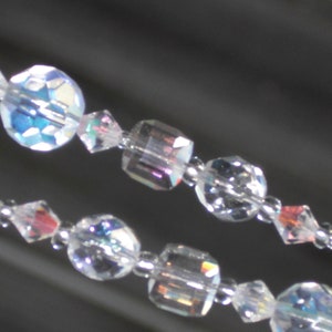 May include: Close-up of two strands of clear, iridescent crystal beads. The beads are various shapes, including round and faceted, with a shimmering, rainbow effect. The beads are strung on a thin, silver-colored wire.