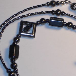May include: Black eyeglass chain with a variety of dark beads. The chain features small, round beads, rectangular beads, and square beads. The ends have loops to attach to glasses. The chain is designed to be worn around the neck.