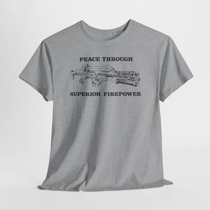 Könnte beinhalten: Ein graues, meliertes Kurzarm-T-Shirt mit einer schwarzen Strichzeichnung eines Gewehrs in der Mitte der Brust. Über dem Gewehr steht der Text PEACE THROUGH und unter dem Gewehr der Text SUPERIOR FIREPOWER in einer fetten, schwarzen Serifenschrift.