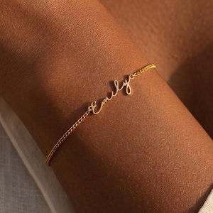 Puede incluir: Una delicada pulsera dorada con el nombre "Emily" en escritura cursiva. La pulsera está en una cadena delgada y se usa en una muñeca. El fondo es una camisa blanca.