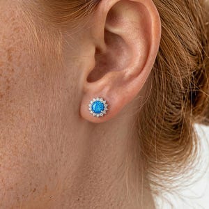 May include: A close-up of an ear with a blue and white stud earring. The earring features a round, light blue centre stone encircled by small, white accent stones. The earring is set in a silver-coloured metal.