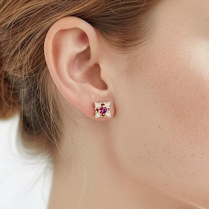 May include: A close-up of a gold square stud earring with a central red gemstone and surrounding smaller white gemstones. The earring is set against the backdrop of a person's ear and face, highlighting the jewelry's design and color.
