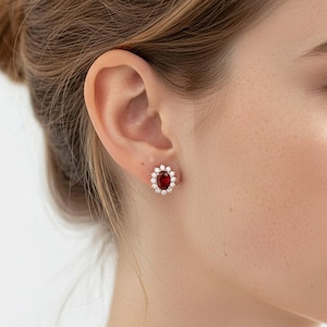 以下が含まれることがあります： 中央に楕円形の深い赤色の宝石があり、その周りを小さな透明な宝石が囲むスタッドピアス。 イヤリングは金色の金属にセットされています。 人の耳に表示されています。