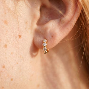 Può includere: Un orecchino a cerchio dorato con quattro pietre preziose rotonde e trasparenti. L'orecchino è indossato sull'orecchio, mettendo in risalto le pietre scintillanti e il tono caldo dell'oro. Il design è semplice ed elegante.