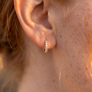 May include: A small gold hoop earring featuring a row of clear, round-cut stones set along the front surface. The hoop is circular in shape and has a polished metallic finish, with the stones catching the light to create a bright, reflective appearance.