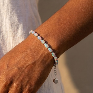 May include: A silver-toned bracelet featuring a series of oval-shaped iridescent opals set in metal prongs. The bracelet includes a delicate chain extension that terminates in a small, flat, heart-shaped charm dangling from the end of the clasp.
