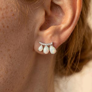 Puede incluir: Primer plano de un pendiente de plata con tres piedras de ópalo en forma de pera. El pendiente tiene una barra curva con pequeñas piedras transparentes sobre las gotas de ópalo. El pendiente se lleva en una oreja.