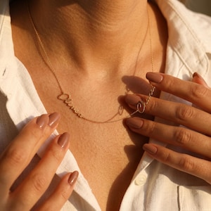Può includere: Una delicata collana color oro con il nome "Oliver" scritto in modo elegante. La collana è indossata su una camicia bianca. Il nome è scritto in un carattere corsivo. La collana è tenuta dalle mani di una persona.