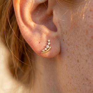May include: A gold ear climber earring featuring a curved, textured metal base set with four round, clear, faceted stones that graduate in size, following the natural contour of the earlobe in a delicate, ascending arrangement.