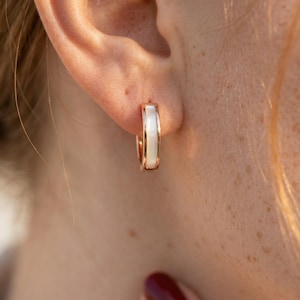 May include: A close-up of a gold-colored hoop earring with a white, rectangular inlay. The earring is worn on an ear, with the focus on the jewelry's design and the contrast between the gold and white elements.