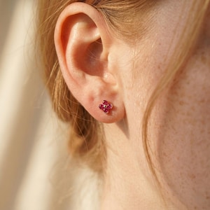May include: A close-up of a person's ear with a small, four-petal flower-shaped earring. The earring features small, bright red gemstones set in a gold-colored metal. The skin around the ear is fair-toned.