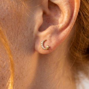 May include: A gold stud earring shaped like a crescent moon and a small star. The crescent moon is encrusted with small, clear, sparkling stones, while the star is a solid gold finish. The earring is positioned on the lower earlobe.