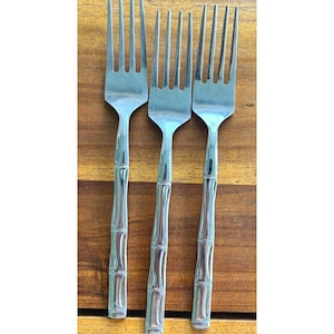 May include: Three stainless steel forks with bamboo-style handles. The forks are displayed vertically against a wooden background, highlighting their tines and the textured handles.