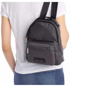 Puede incluir: Una mochila negra de nailon con un compartimento principal con cremallera y un bolsillo frontal. La mochila tiene correas ajustables para los hombros y un asa superior. El nombre de la marca "MARC JACOBS" es visible en el bolsillo frontal.
