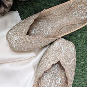 May include: A pair of flat slip-on shoes featuring a metallic gold fabric base with intricate silver thread embroidery in swirling floral patterns. The shoes have a pointed toe design and a scalloped edge along the opening, with matching floral embroidery on the inner sole.