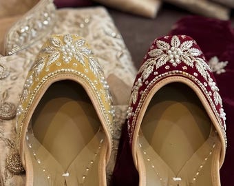 May include: A pair of ornate, embroidered slip-on shoes in yellow and burgundy. The shoes feature intricate floral designs with pearl accents. Traditional formal wear is visible in the background.