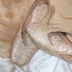 May include: A pair of blush-colored, embroidered flats. The shoes feature intricate silver-colored floral embroidery across the satin fabric. The flats are displayed on a white and gold-toned fabric background.