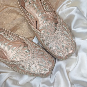 May include: A pair of blush-colored, embroidered flats with intricate silver floral patterns. The shoes are made of a satin-like material and are displayed on a white and gold fabric background. The flats have a pointed toe design.