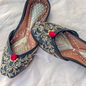 May include: A pair of black flat shoes featuring intricate gold floral embroidery across the upper surface. Each shoe is accented with a small, bright red pom-pom at the center. The interior lining is tan with a metallic gold diamond pattern.