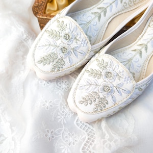 Può includere: Un paio di ballerine bianche con ricami floreali argentati e azzurri. Le scarpe sono esposte su un tessuto di pizzo bianco, che mette in risalto le cuciture e il design dettagliati. Le ballerine hanno una punta arrotondata e un bordo dorato.