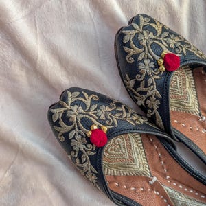 May include: A pair of black flats with intricate gold embroidery and a small red pom-pom accent. The shoes feature a leather interior with a tan and gold design. The flats are a traditional style, likely for special occasions.
