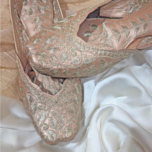 May include: A pair of blush-colored, embroidered slip-on shoes. The shoes feature intricate silver-colored floral embroidery across the top and sides. The shoes are made of a soft, satin-like material and are displayed on a white satin surface.