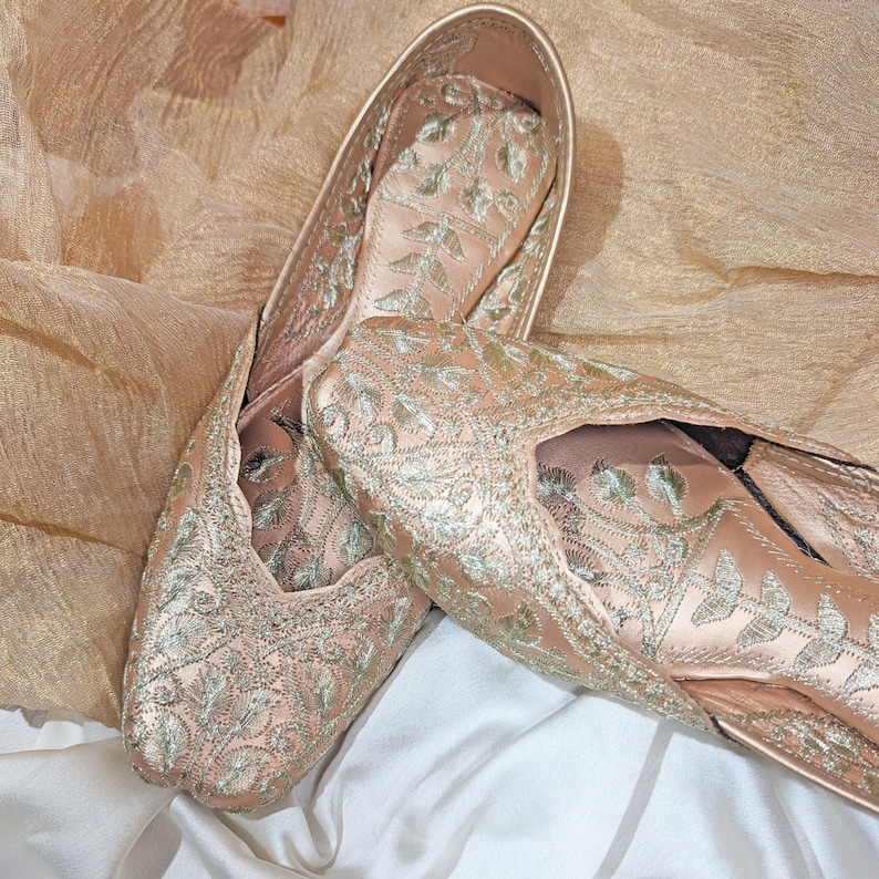 May include: A pair of blush-colored, embroidered flats. The shoes feature intricate silver-colored floral embroidery across the satin fabric. The flats are displayed on a white and gold-toned fabric background.