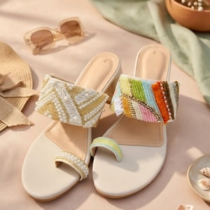 May include: Cream sandals with decorative beading. One sandal has gold and pearl detailing, the other a colourful beaded strap. The image also includes sunglasses and a straw hat, suggesting a summer theme.