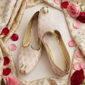 Puede incluir: Un par de zapatos estampados de color crema con ribetes dorados. Los zapatos están rodeados de tela color crema con bordados florales dorados, pétalos de rosa y rosas rosas y rojas. Los zapatos tienen punta y tacón bajo.