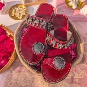 Könnte beinhalten: Rote Ledersandalen mit metallenen Kamelmotiven an den Riemen und kreisförmigen Verzierungen. Die Sandalen werden in einem Weidenkorb präsentiert, umgeben von rosa Rosenblättern. Ein Paar Sandalen.